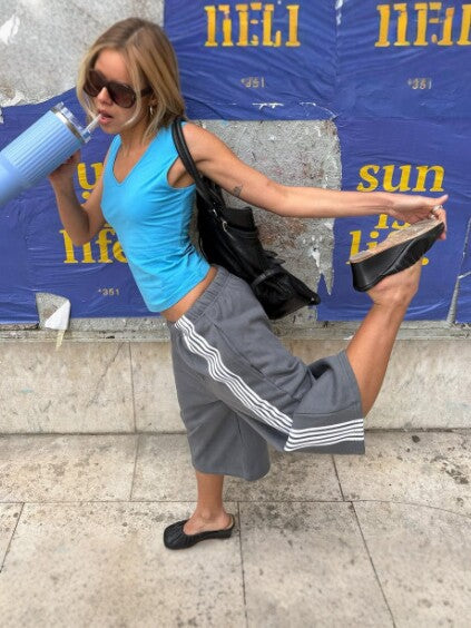 Woman in gray striped wide-leg pants lifting one leg in a dance pose, blue tank top and sunglasses