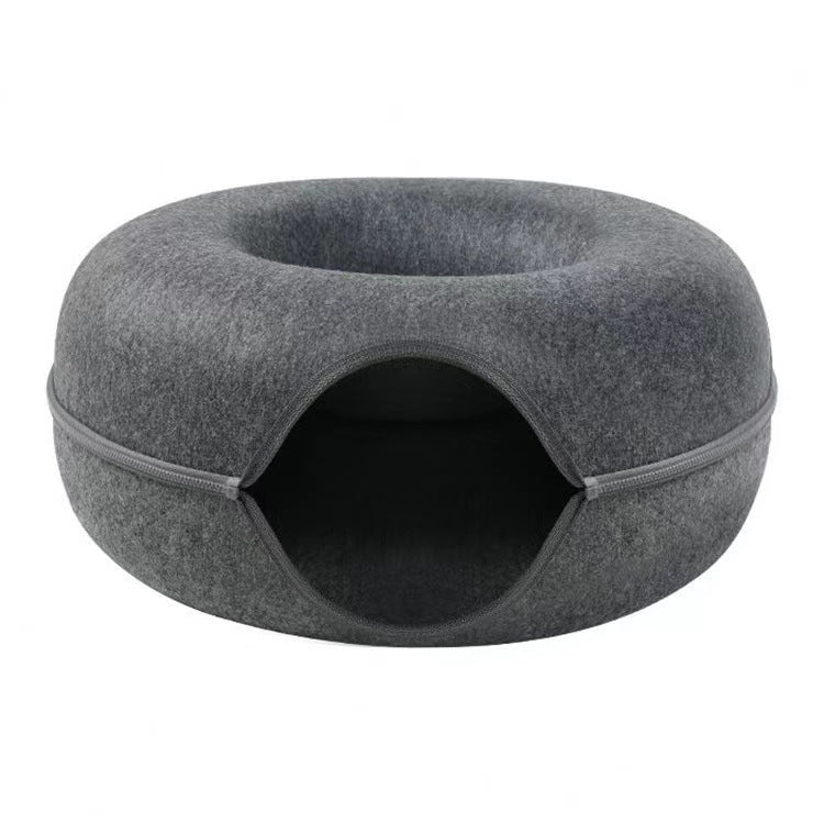 Two cats sharing a large beige felt donut tunnel bed on a wooden floor