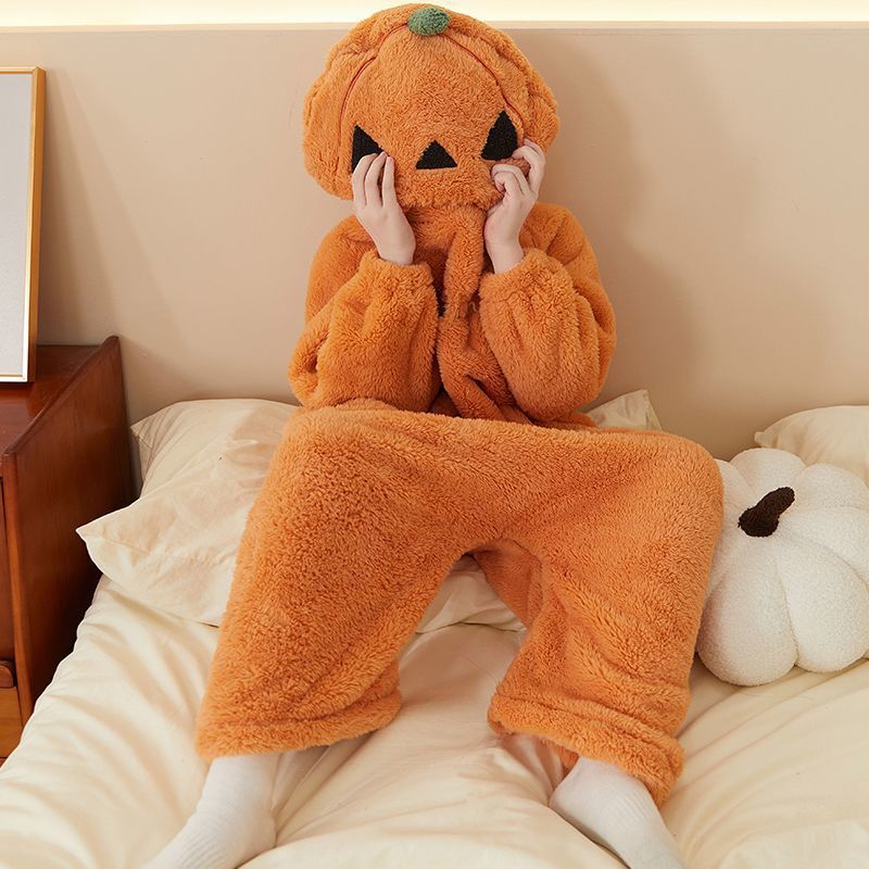 Person lounging on a bed in an orange fleece pumpkin onesie with pumpkin hood beside a white pumpkin cushion