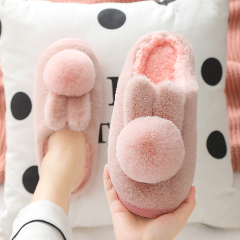 Soft pink plush bunny slippers with pom pom tails, one held in a hand and one worn on a foot, pink variant