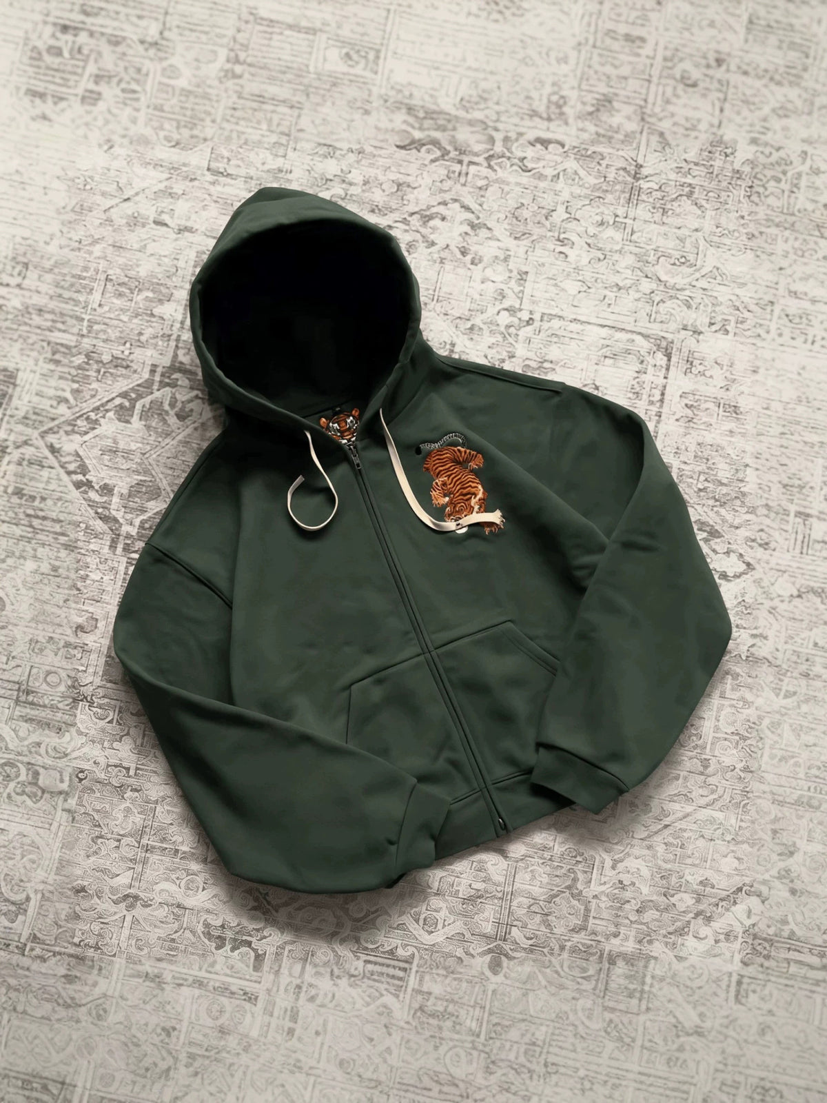 Green zip-up hooded sweatshirt flat lay with embroidered orange tiger on the chest and a cream drawcord