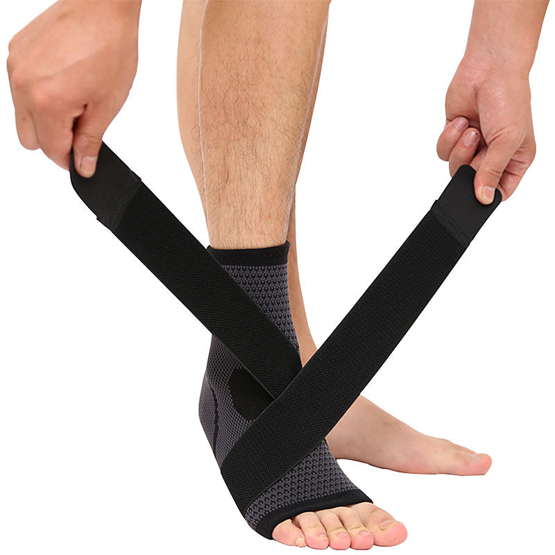 Black and grey knit ankle brace with a long compression strap crossing the foot, held by two hands on a white background