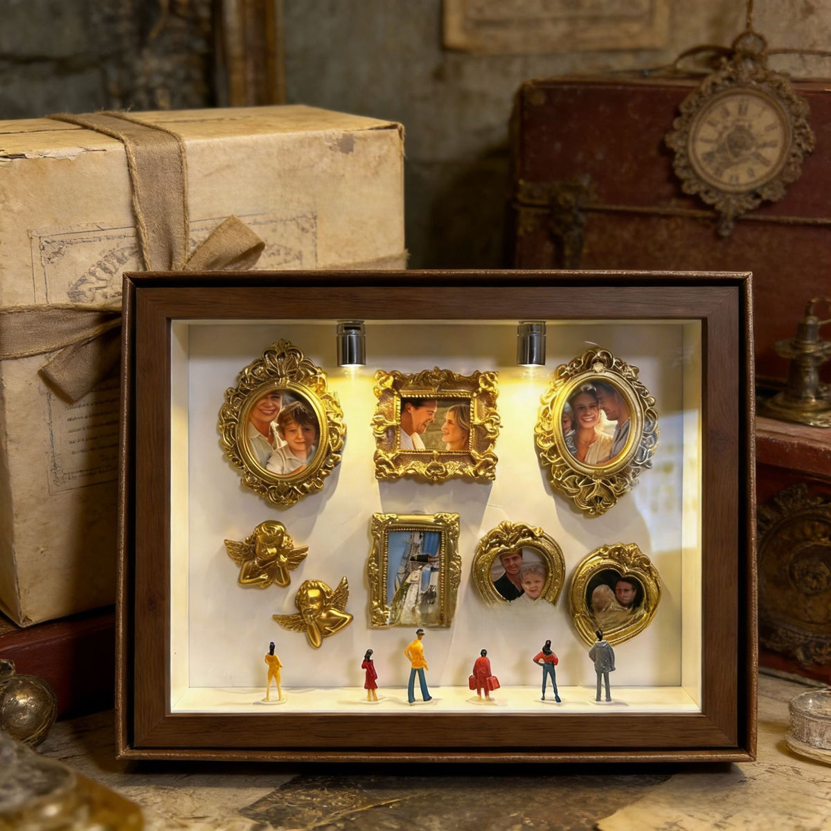 Dark brown shadow box set with gold mini photo frames, cherubs and visitor figurines lit by two lights on a vintage table