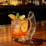 Clear novelty glass mug with a cheeky sculpted handle holding an iced cocktail with lemon on a bar