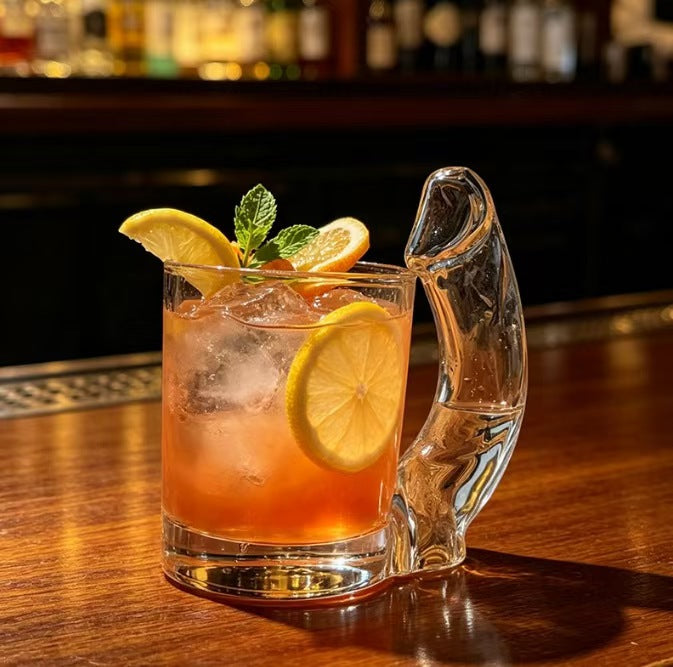 Clear novelty glass mug with a cheeky sculpted handle holding an iced cocktail with lemon on a bar