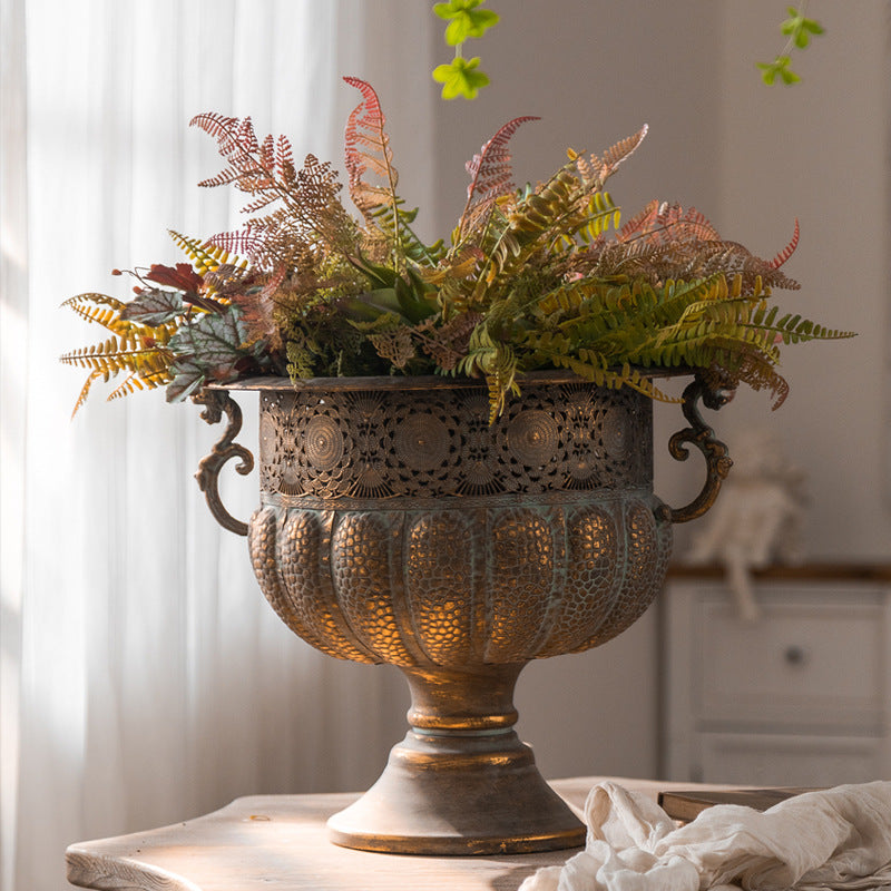 Vintage iron pedestal urn with openwork band filled with ferns beside sheer curtains on a tabletop