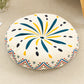 Round cushion with a teal and yellow burst print on a cream top and a zigzag border, on a beige rug
