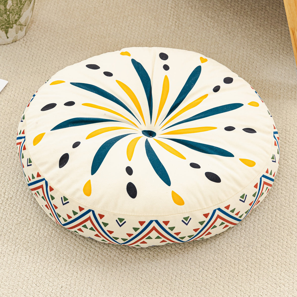 Round cushion with a teal and yellow burst print on a cream top and a zigzag border, on a beige rug