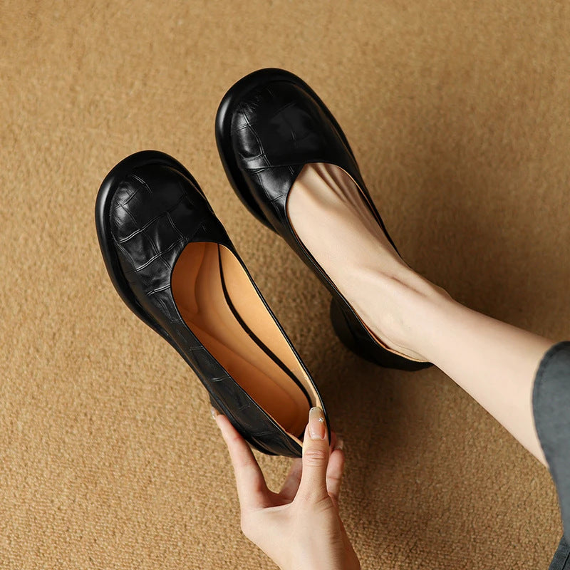 Black embossed leather round toe slip-on shoes, one held to show the tan insole, the other worn on a foot