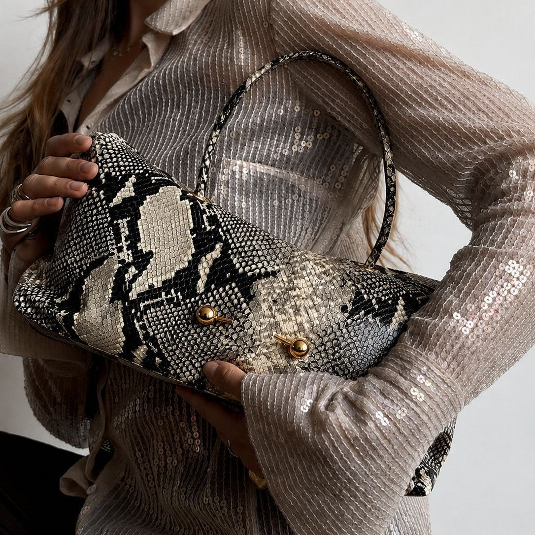 Woman holding a snake print shoulder bag under her arm over a sequin top, gold ball studs visible