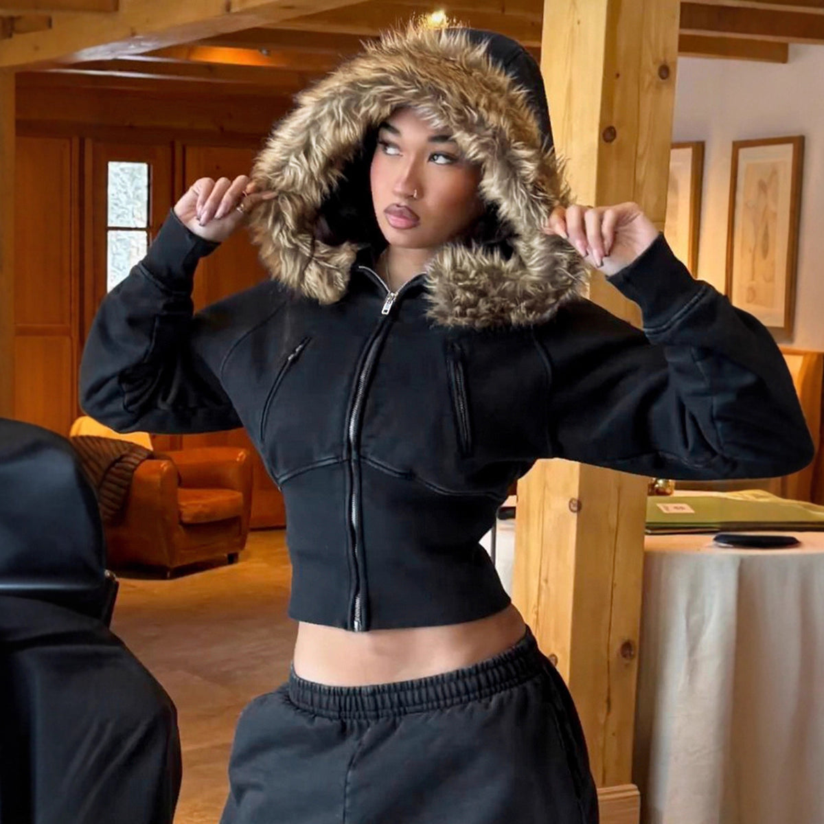 Model in a black cropped fur trimmed hooded zip jacket with a tan plush fur hood raised around the face