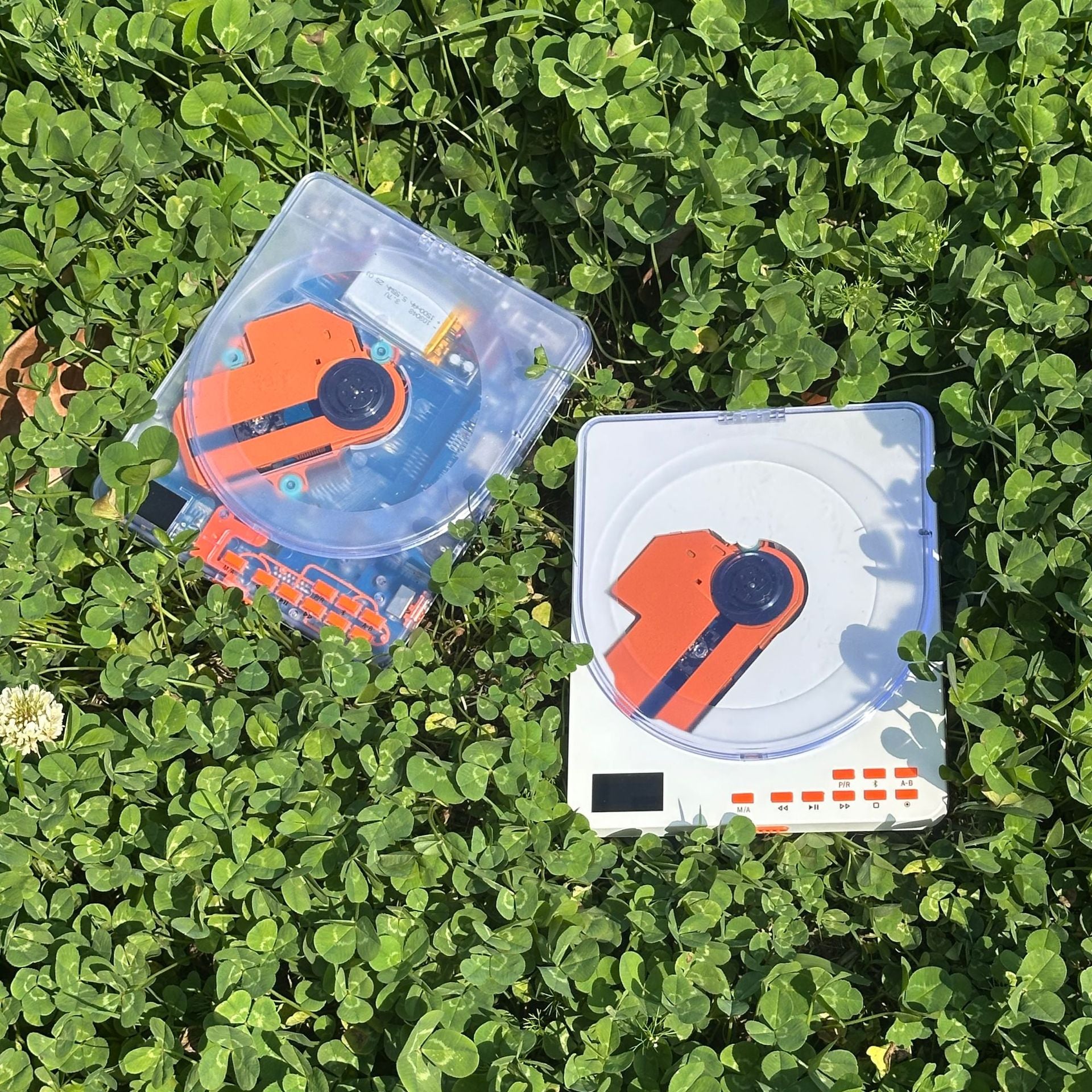 Two portable CD players in clover, one transparent shell and one white shell