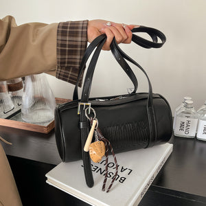 Hand holding a black cylinder shoulder bag by two long straps with a buckle strap and a baseball glove charm