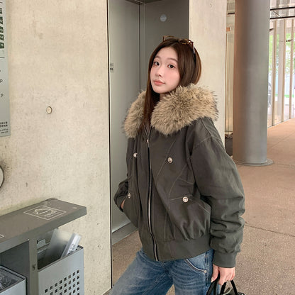 Woman in a dark gray green hooded bomber jacket with vegan fur collar and metal snap details by a concrete wall