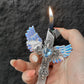 Hand holding the lit angel wings cross lighter with the flame rising and a blue heart rhinestone at the centre