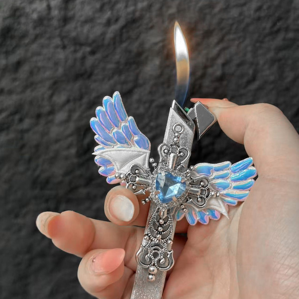 Hand holding the lit angel wings cross lighter with the flame rising and a blue heart rhinestone at the centre