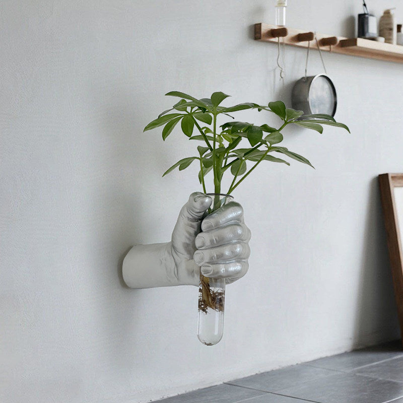 Metallic hand shaped wall vase holding a glass tube with a leafy stem against a plain wall
