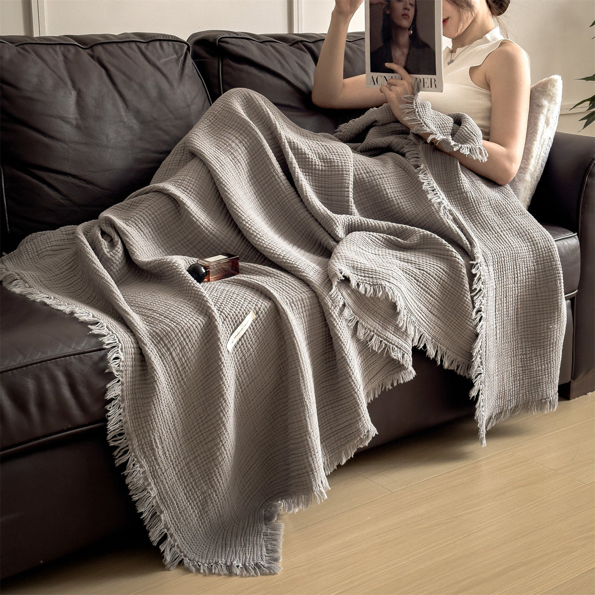 Mist Gray cotton gauze throw blanket with tassel fringe, draped over a seated woman on a dark leather sofa, view 2 of 4