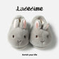 Pair of gray bunny plush slippers with pink inner ears seen from above on a light panel with script lettering, view 2 of 3