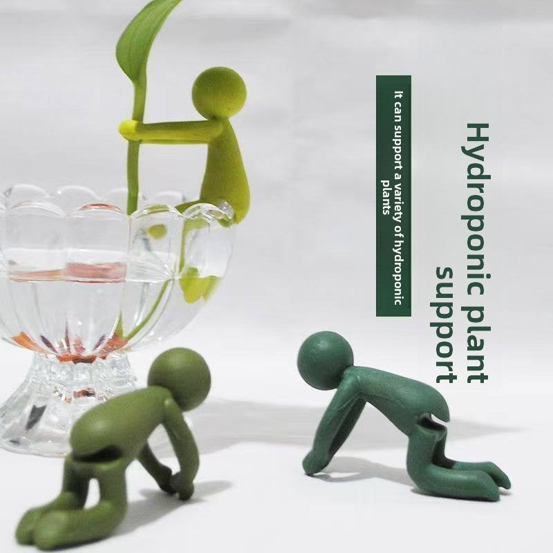 Green figures climbing a stem and crawling beside a glass vase, caption Hydroponic plant support