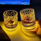 Two lit whiskey tumblers on a desk, one held, amber glow spreading across the surface