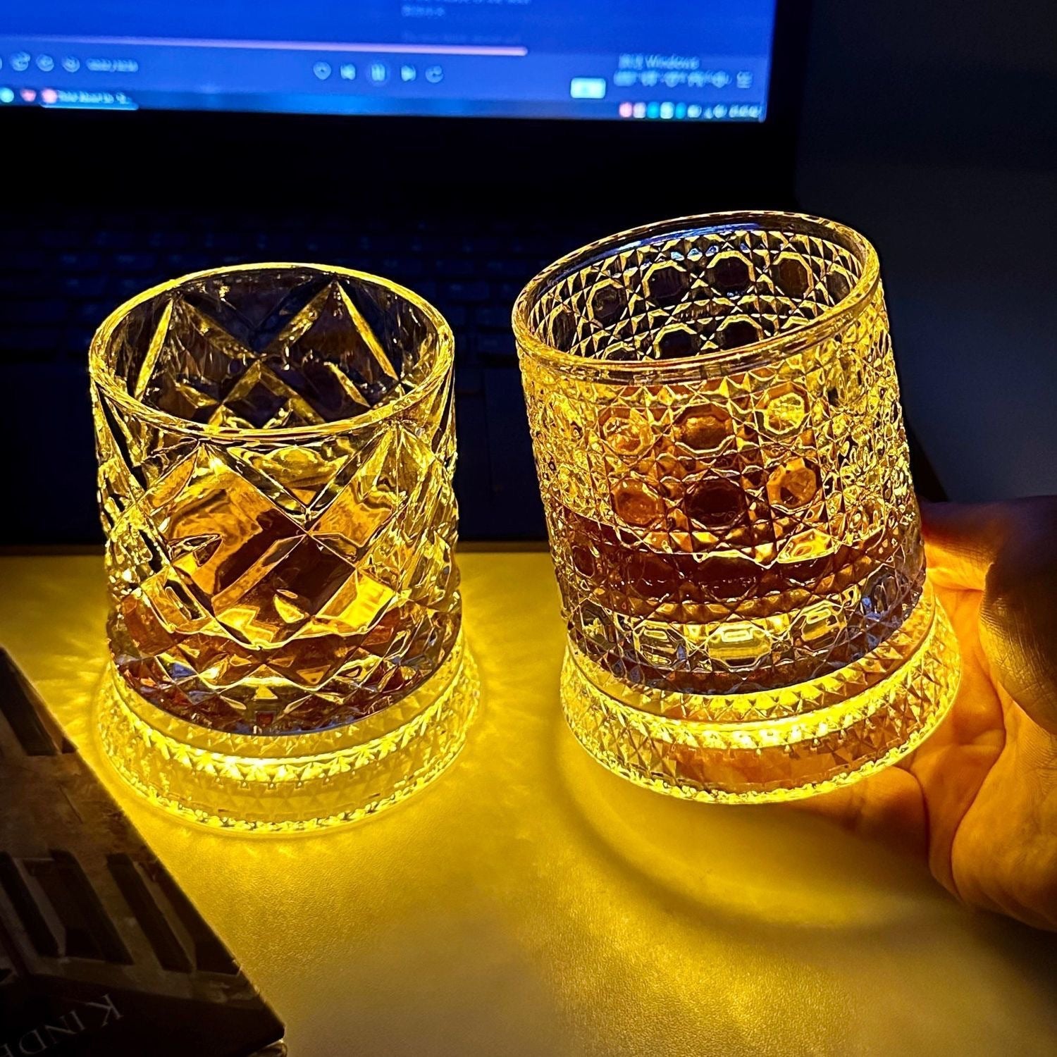 Two lit whiskey tumblers on a desk, one held, amber glow spreading across the surface