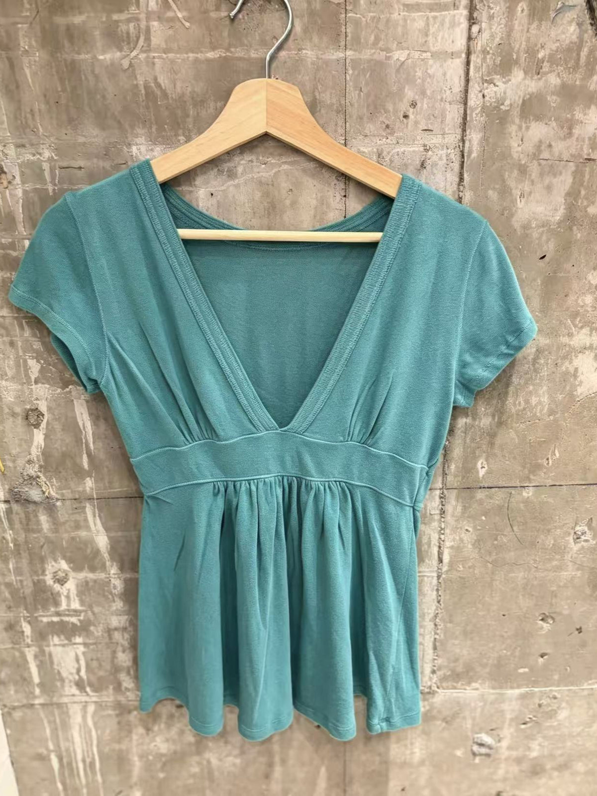 Teal short-sleeve deep V-neck top with an empire waist band and gathered hem on a wooden hanger