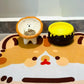 Lion bowl of kibble beside a brown honey jar bowl with yellow drips on a cat face patterned mat