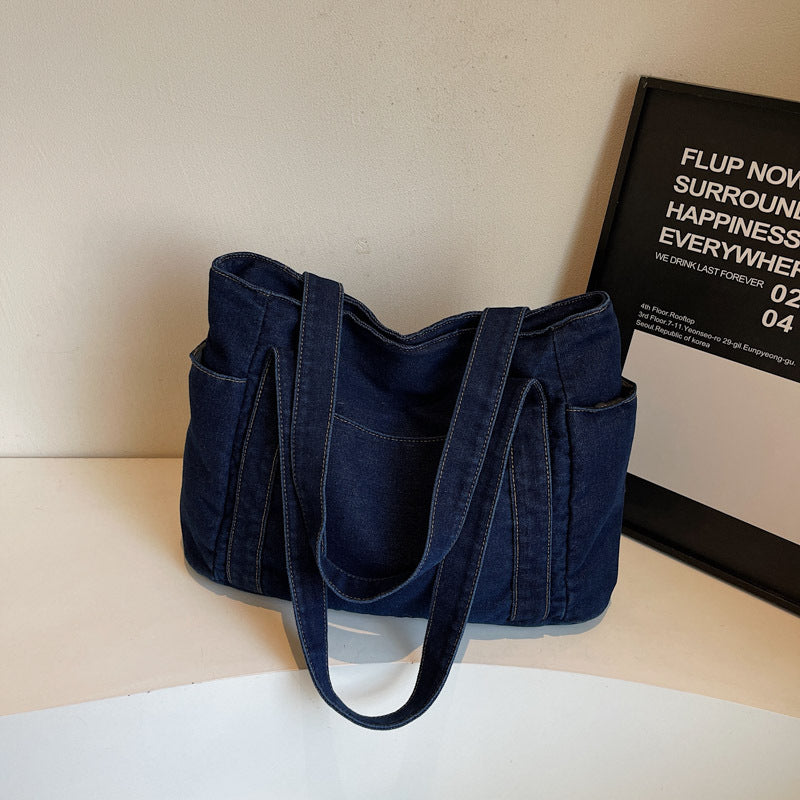 Dark blue denim shoulder tote beside a framed poster, front patch pocket and twin shoulder straps visible