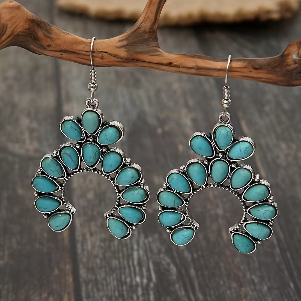 Pair of crescent drop earrings with turquoise colored stones hanging from a branch above dark wood planks