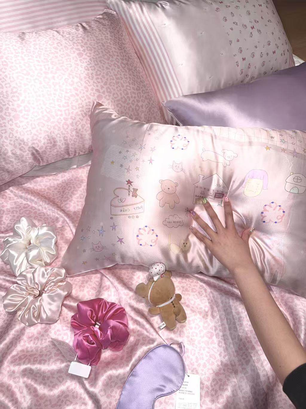 Printed and plain pink and lilac silk pillowcases with scrunchies and a sleep mask on a pink bed