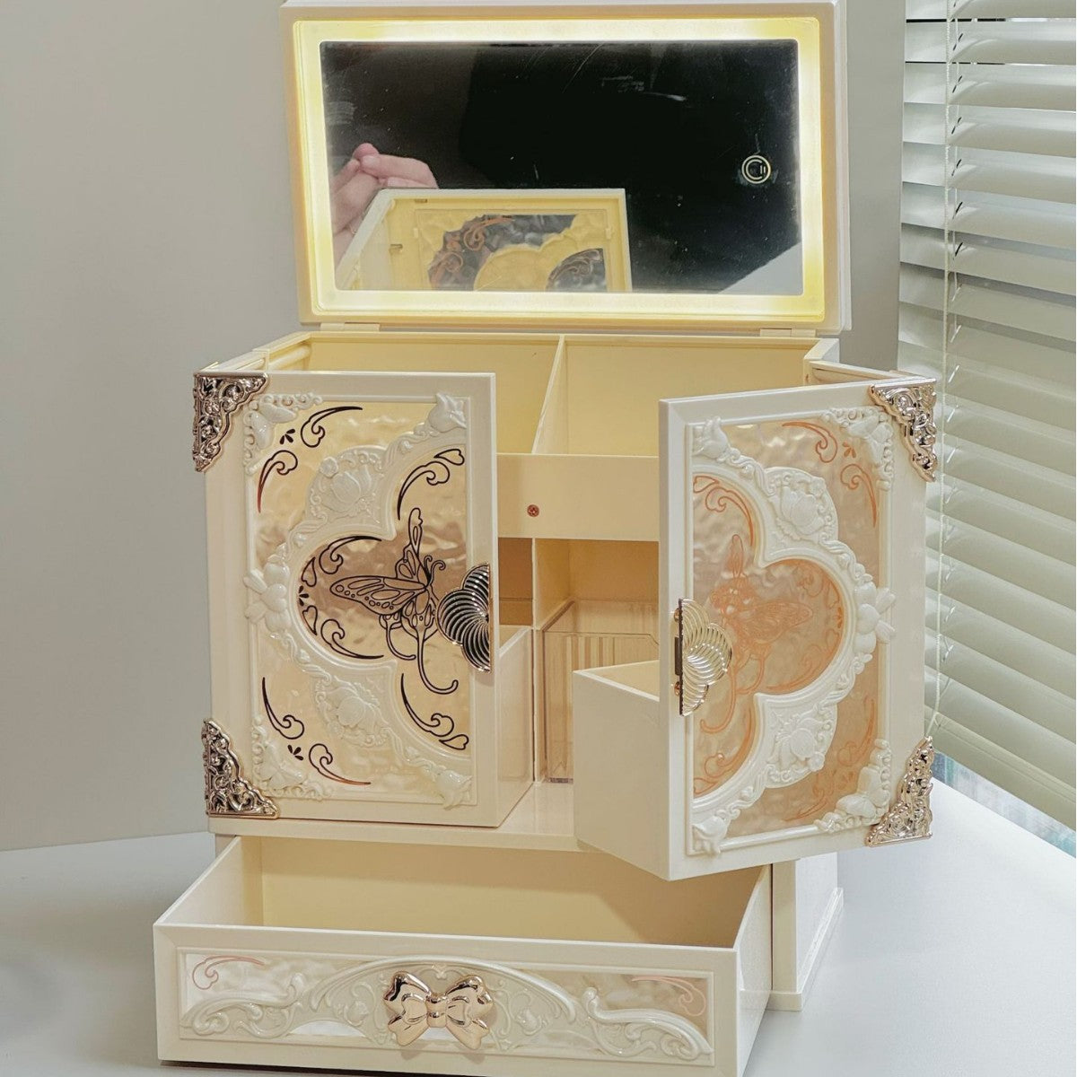 Open ivory makeup organizer cabinet with a warm lit mirror in the raised lid, both doors open and the drawer pulled out