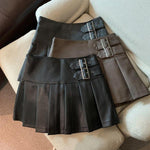 Three pleated mini skirts with buckle straps in black, brown and dark gray laid on a cream sofa