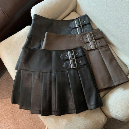 Three pleated mini skirts with buckle straps in black, brown and dark gray laid on a cream sofa
