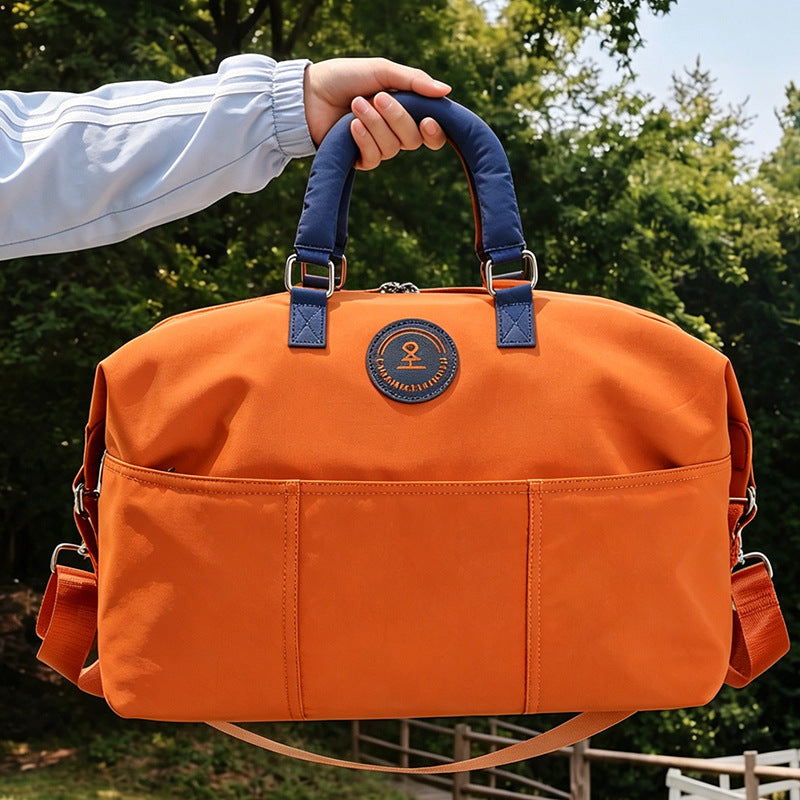 Hand holding an orange duffel travel bag by its navy blue padded handle outdoors, with front slip pockets