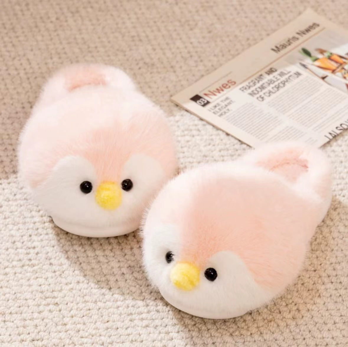 Pink plush penguin slippers with yellow beaks and black eyes on a beige rug beside a newspaper