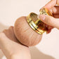 Hand holding gold handle mushroom powder brush against a pale background