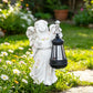 White resin angel statue with pink rose accents holding an unlit black lantern on a sunny garden lawn