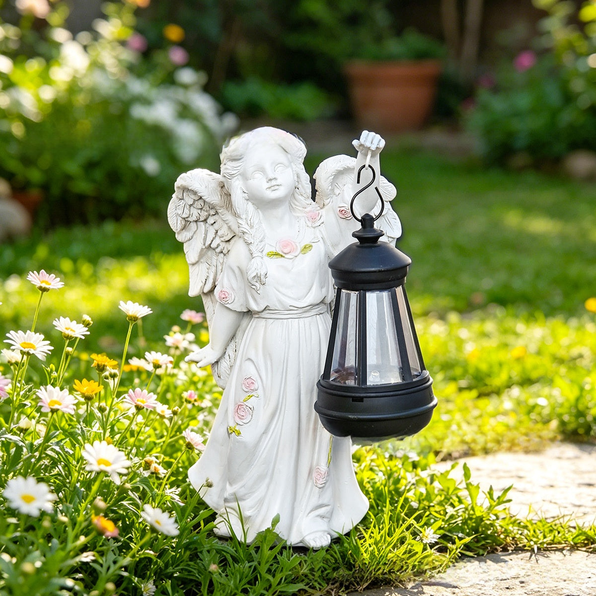 White resin angel statue with pink rose accents holding an unlit black lantern on a sunny garden lawn