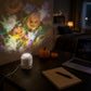 White projector lamp with a cable on a dark desk beside a laptop, throwing pumpkin and ghost patterns on the wall
