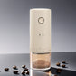 Cream electric coffee grinder with a round front button and numbered grind ring, coffee beans scattered on a dark surface