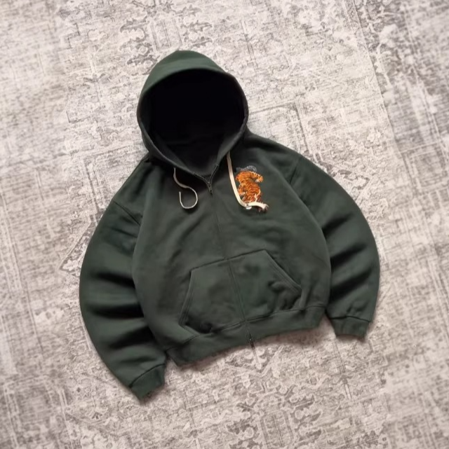 Green boxy zip-up hoodie with front pocket and ribbed cuffs lying on a patterned rug with a tiger embroidery
