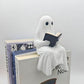 Reading ghost statue sitting on the corner of a pile of large books with its legs hanging over