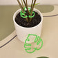 Green monstera leaf plant clip around stems in a white pot, with a second green leaf clip beside the pot