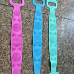 Three silicone back scrubber straps in pink, blue and green showing the fine bristle side on a tiled floor