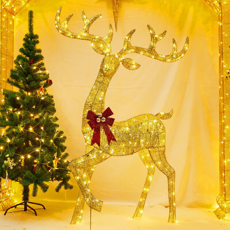 Large golden lit reindeer with a red bow stepping beside a decorated Christmas tree in a warm yellow room