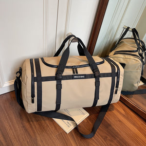 Beige travel duffel bag with black handles and straps standing on a wood floor by a mirror