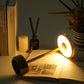 Gold table lamp lit with a warm glow lying on an open book beside a candle and a reed diffuser
