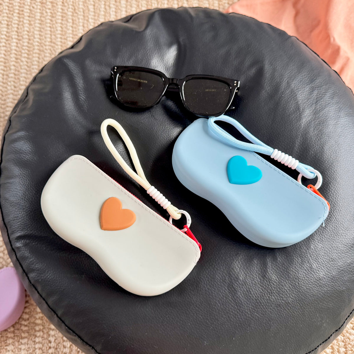 Gray white and blue silicone sunglasses cases with raised hearts and wrist lanyards near sunglasses