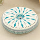 Round cushion with a teal teardrop sunburst and black dots on a cream top with a teal woven border
